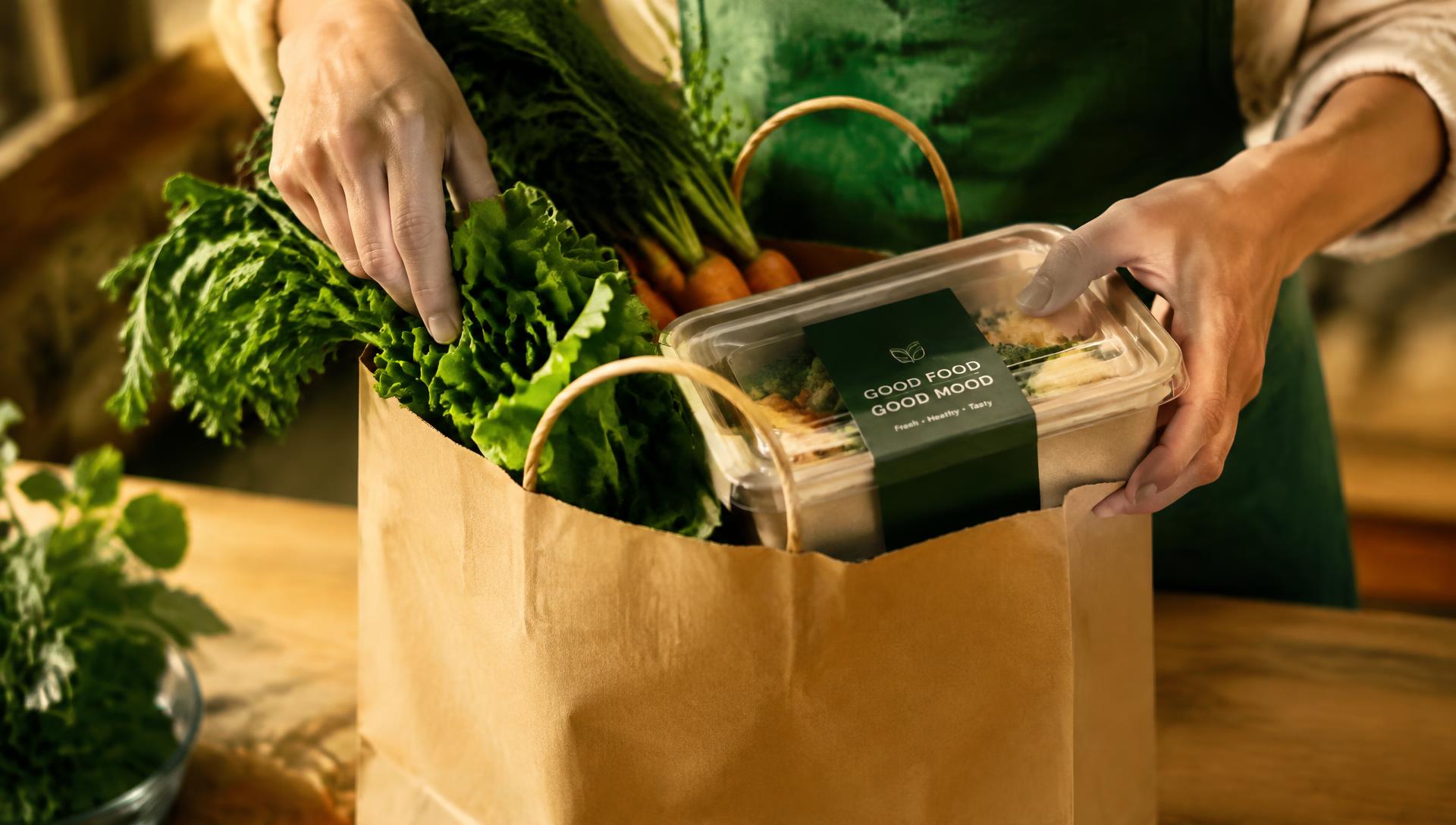 Hands packing fresh vegetables and a meal box into a paper bag